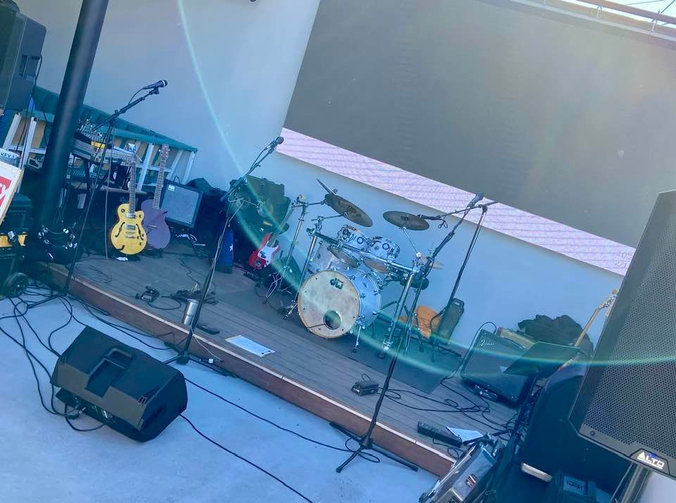 A band's stage setup outdoors, with drums, guitars, and audio equipment. The stage is on a wooden platform, with a white backdrop.
