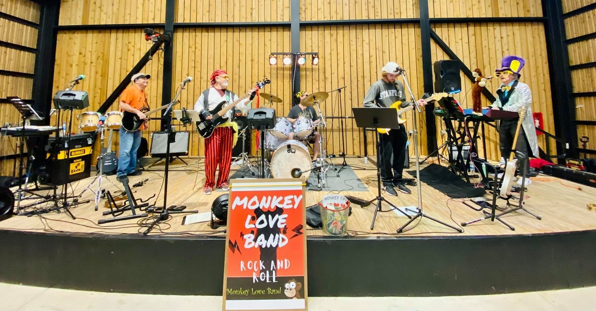 Monkey Love Band performing on a stage with wood paneling. Band members wear colorful outfits and play instruments. A sign promotes their show.