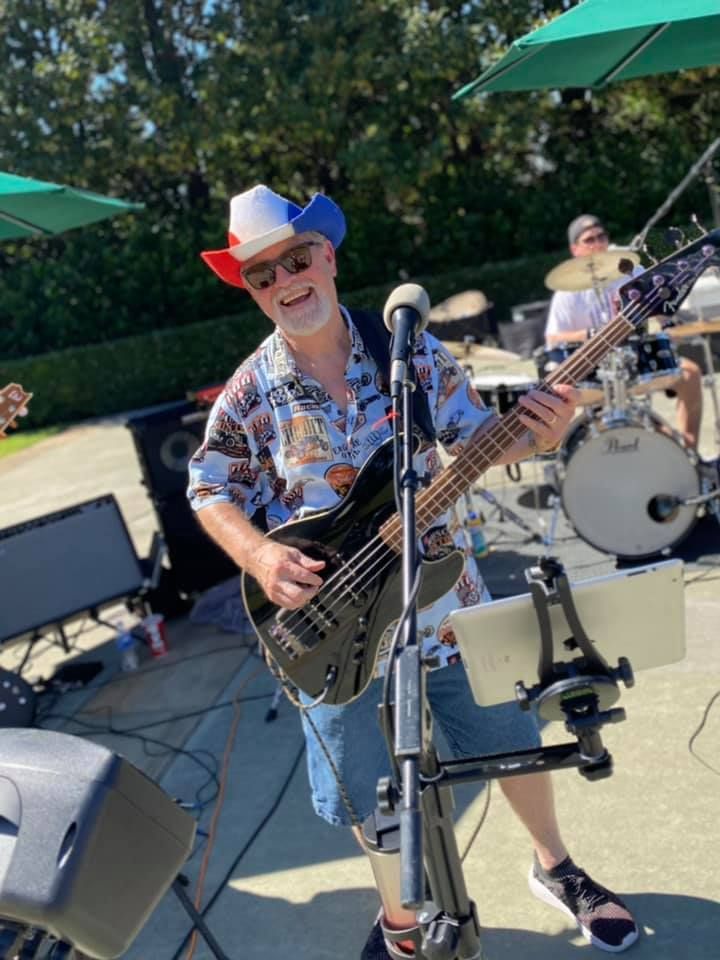 Man playing a black bass guitar outdoors, wearing a patriotic hat and floral shirt, with a drummer in the background.
