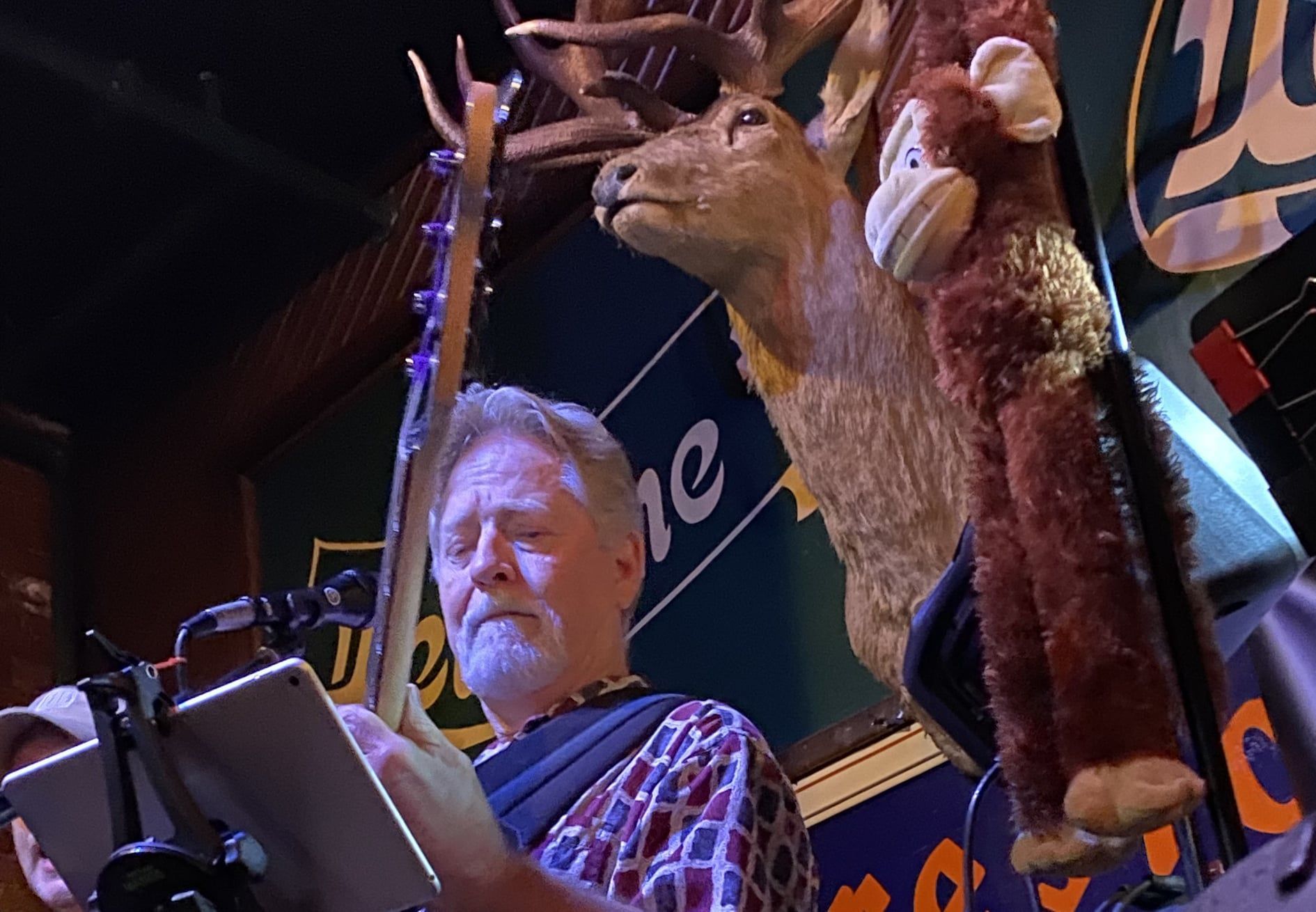 Man with gray beard reading from a tablet at a microphone, in a bar with deer head and stuffed monkey decorations.