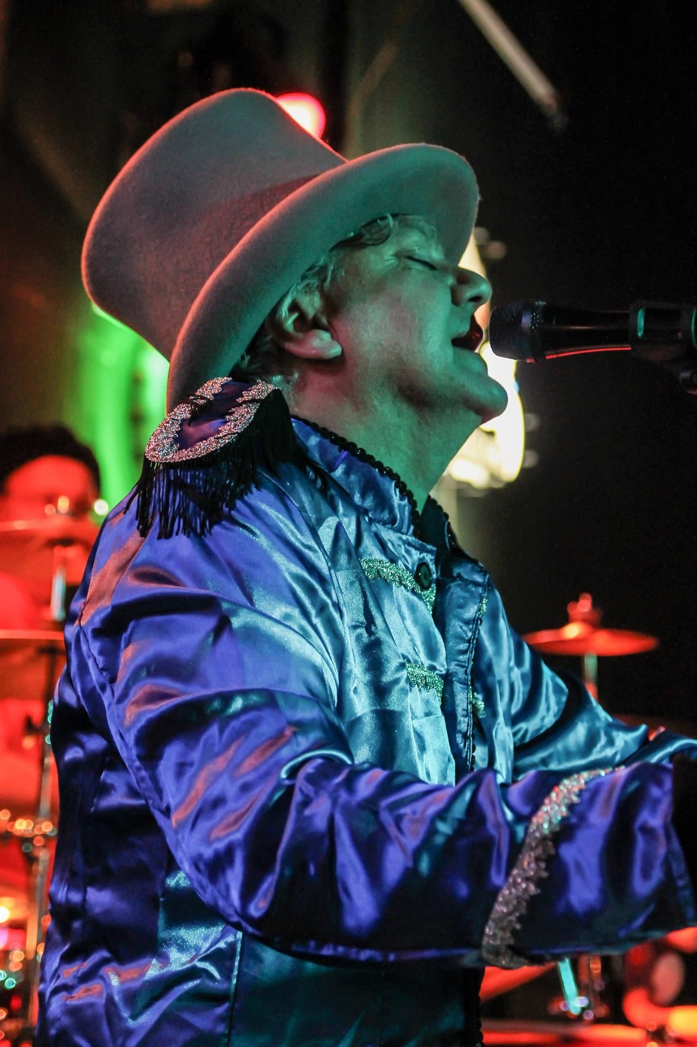 A musician in a shiny blue jacket and top hat sings into a microphone. A drummer is in the background.