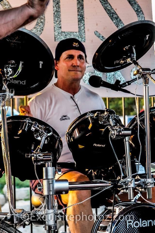 Drummer wearing a cap and white shirt, playing a Roland electronic drum kit outdoors. He smiles, lit by sunlight.