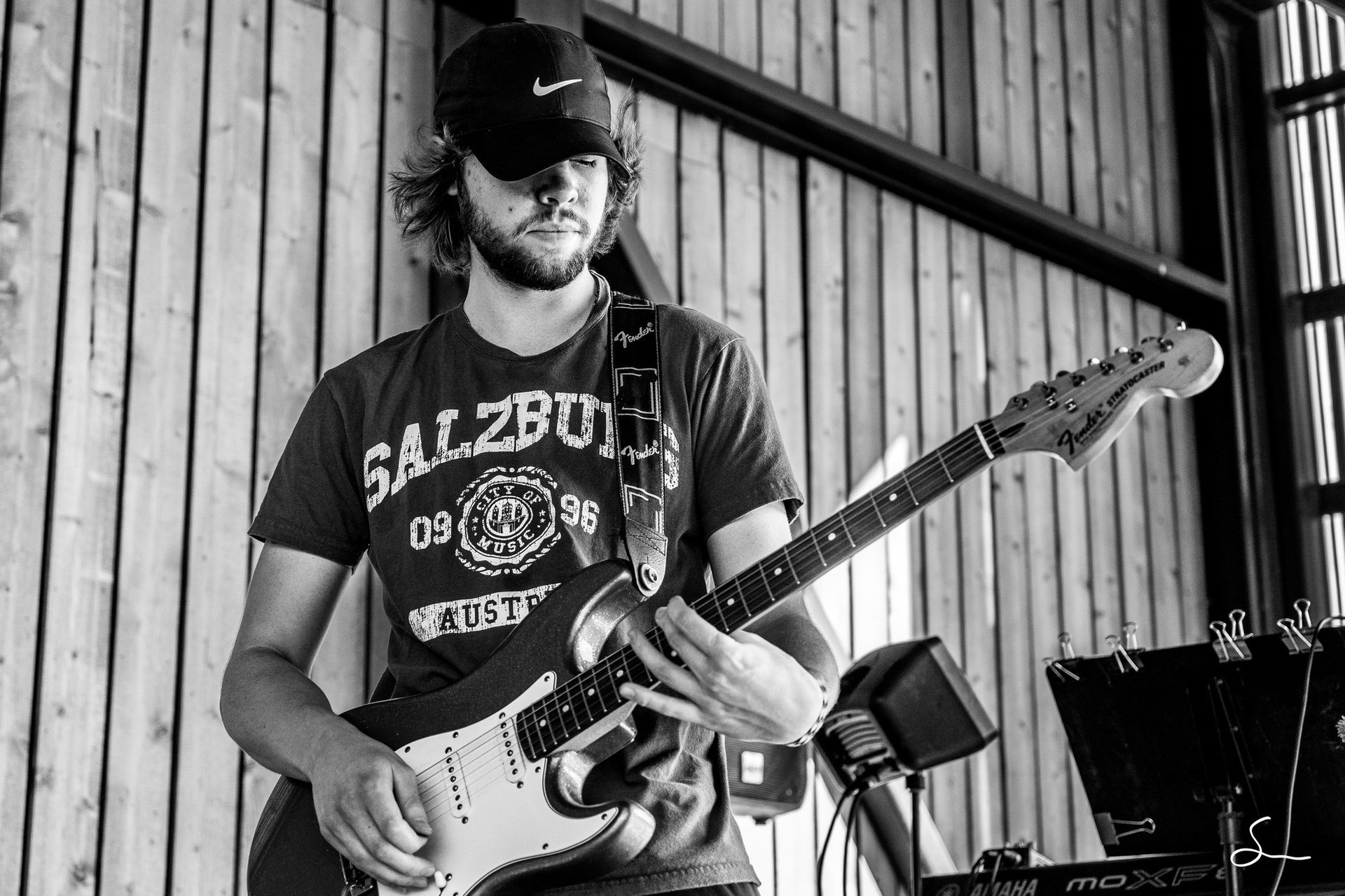 A man with a beard wearing a cap and t-shirt plays an electric guitar in front of a wooden wall.