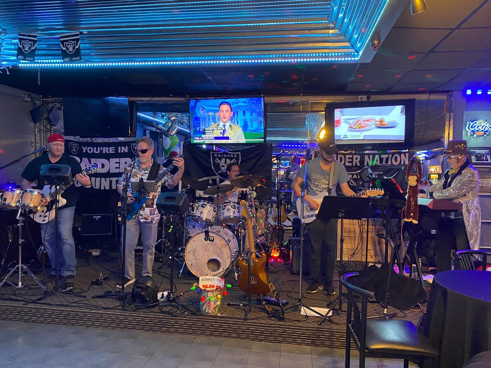 A band performs on a stage in a dimly lit bar. Six musicians play instruments like drums, guitar, and keyboard.