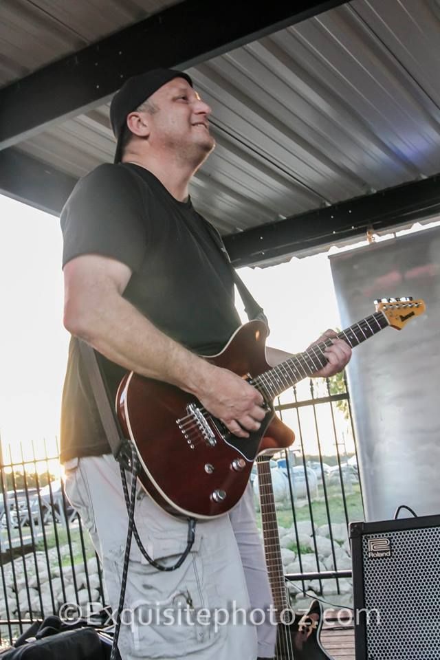 Man playing a brown electric guitar on stage, wearing a black shirt, baseball cap, and light pants. Outdoor setting.