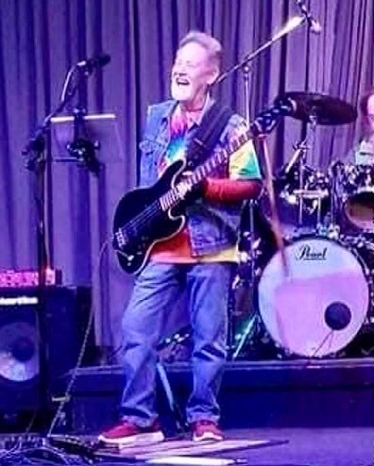 A smiling musician plays a black bass guitar on stage. He wears a tie-dye shirt, denim vest, jeans, and red sneakers, with drums and a speaker visible behind him.