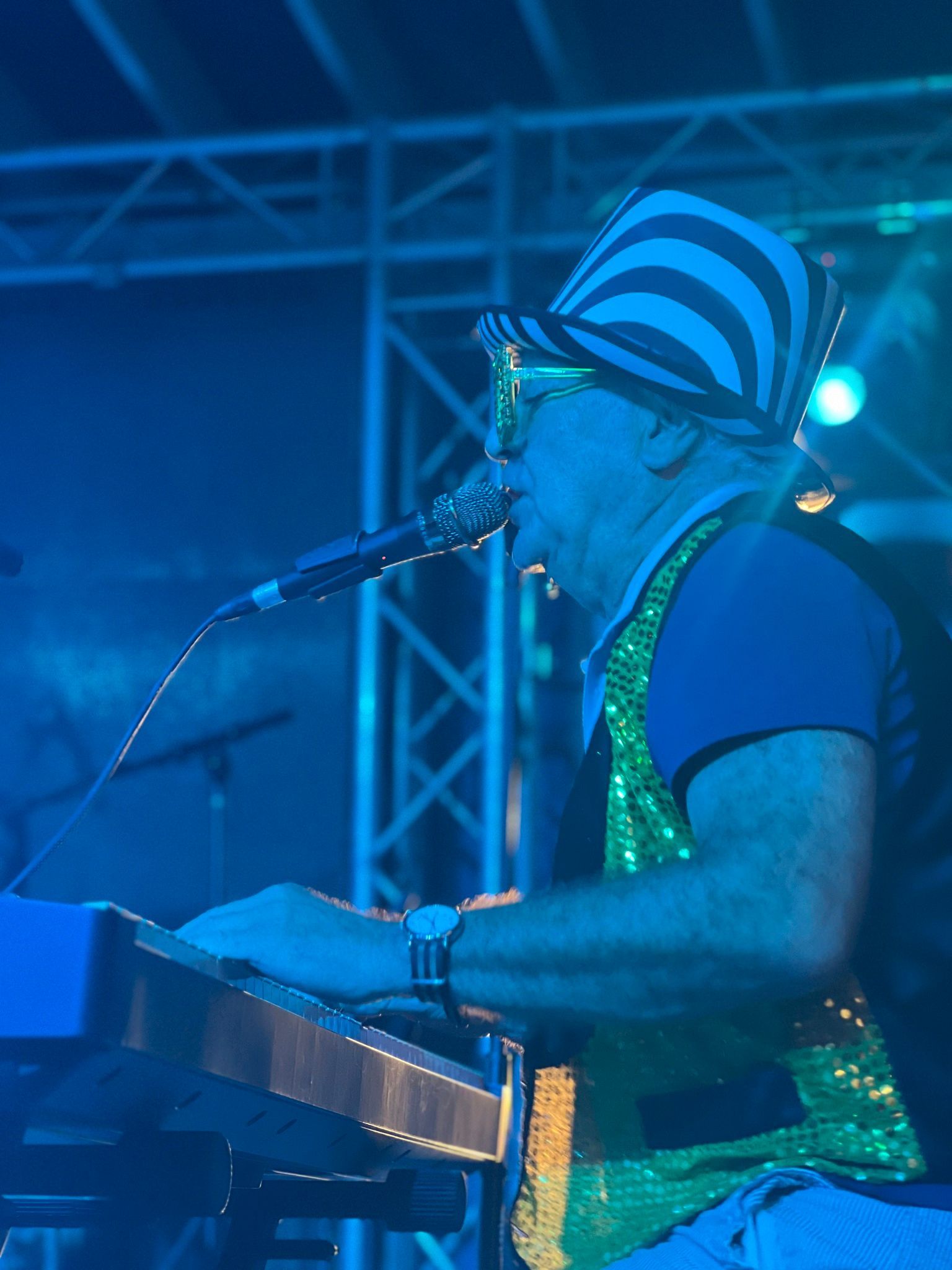 A man in a striped hat and sequined vest plays a keyboard and sings into a microphone on stage, bathed in blue light.