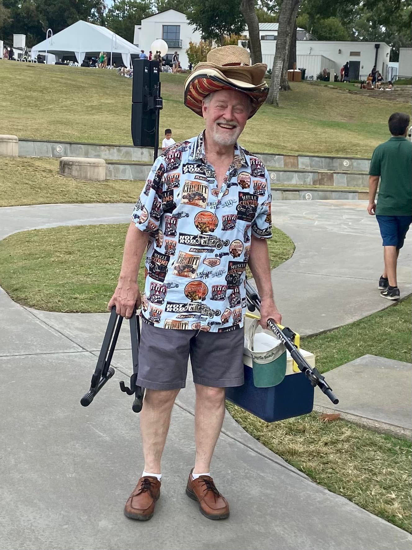 Smiling older man in a cowboy hat and patterned shirt carries a toolbox and tripod on a paved path outdoors.