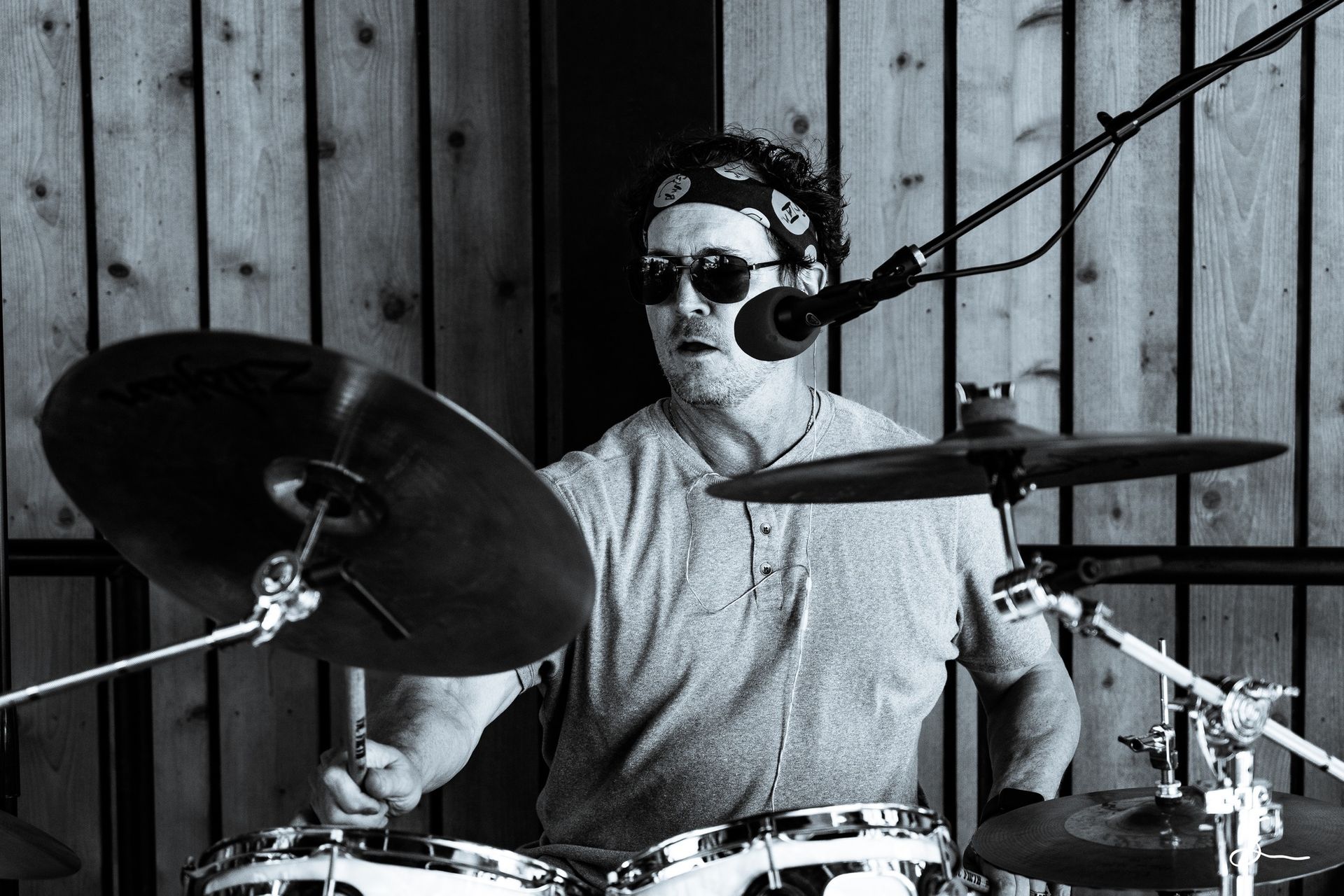 Drummer in sunglasses and a headband, playing a drum set outdoors, with a microphone overhead. Black and white.
