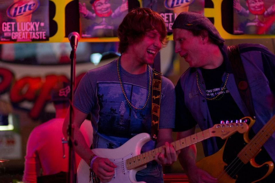Two guitarists on stage, one smiling, one wearing a bandana. Neon lights and Miller Lite signs in the background.