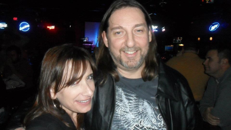 A woman with dark hair and a man with long brown hair smile at the camera in a bar. The man wears a leather jacket.