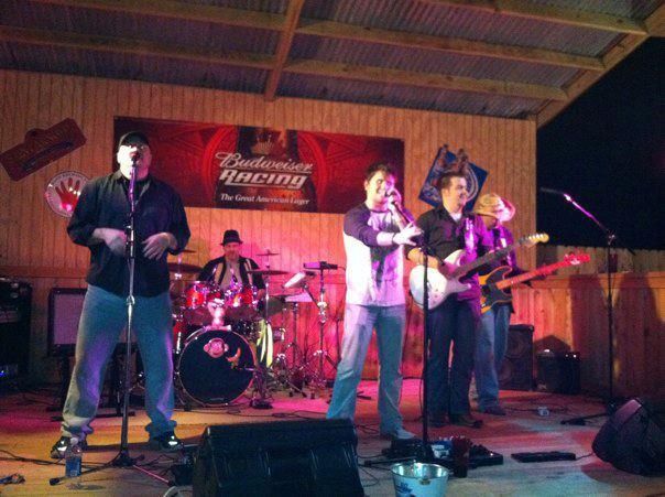 A band of five men performing on stage with microphones and instruments, under dim lighting. The setting appears to be an outdoor venue with a Budweiser sign in the background.