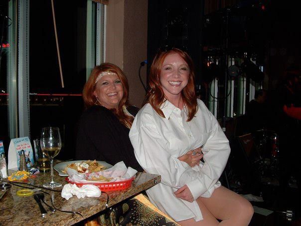 Two smiling women with red hair at a bar. One wears a white shirt and the other a black top, with a red basket of food on the table.