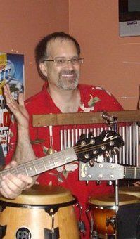 Man with glasses and a red shirt smiles, holding a guitar near conga drums, in what appears to be a music venue.