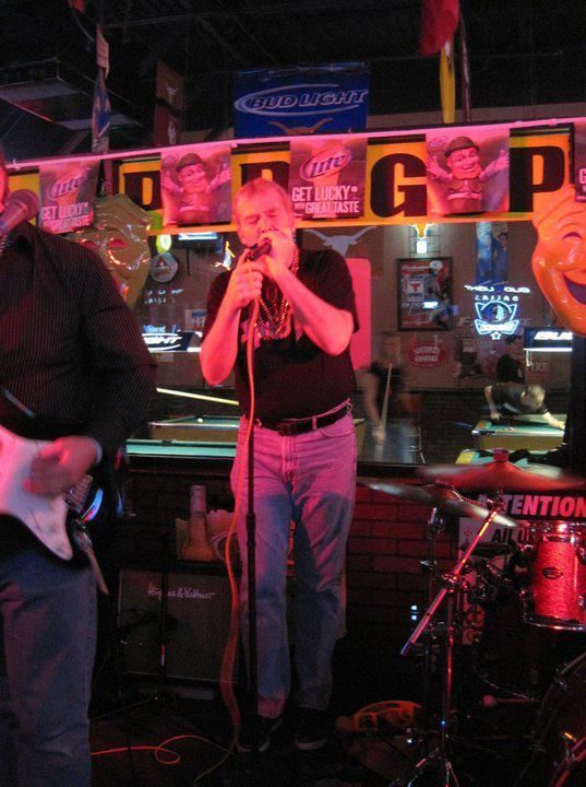 Man singing into a microphone on stage in a dimly lit bar, with a guitar player to his left and drum set on the right.