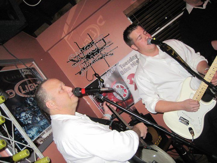 Two men in white shirts performing live music in a bar. One sings into a microphone and plays a drum, the other sings and plays a white electric guitar.