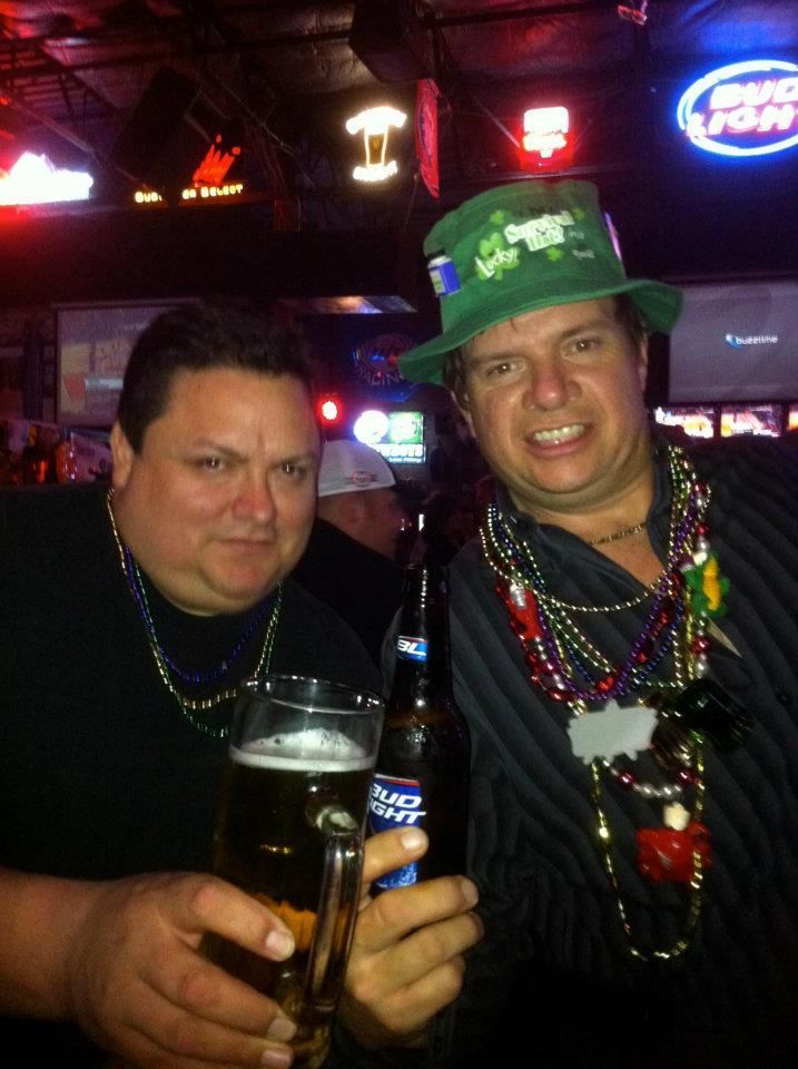 Two men at a bar celebrating, one in a St. Patrick's Day hat, holding beer and bottles.  The setting is dimly lit with neon signs.