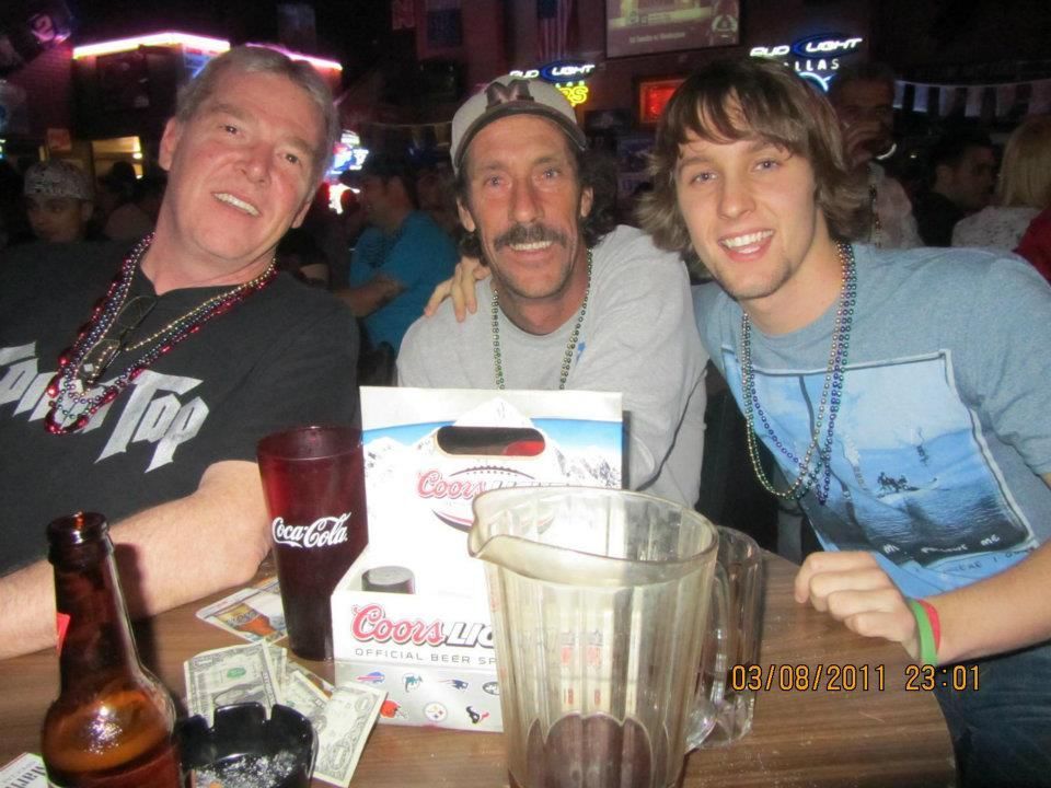 Three men smiling at a bar, two with arms around each other. A table has drinks and money.