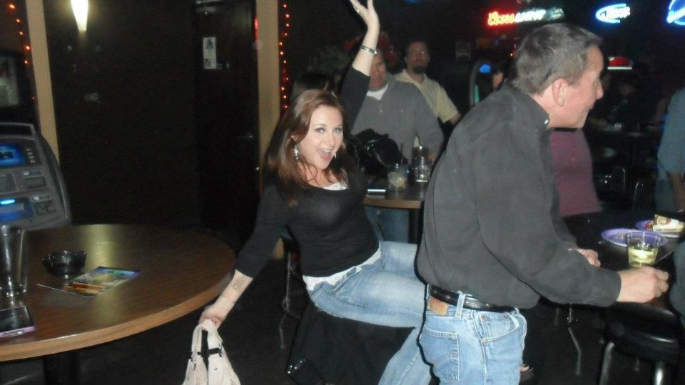 Woman cheering while sitting on a chair, man dancing in a bar. Dimly lit, with bar setting and other people in the background.