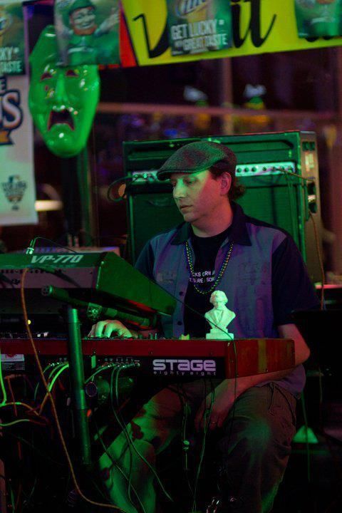 A man plays a red keyboard onstage, wearing a hat and blue shirt. He sits in front of green stage equipment under neon lights.