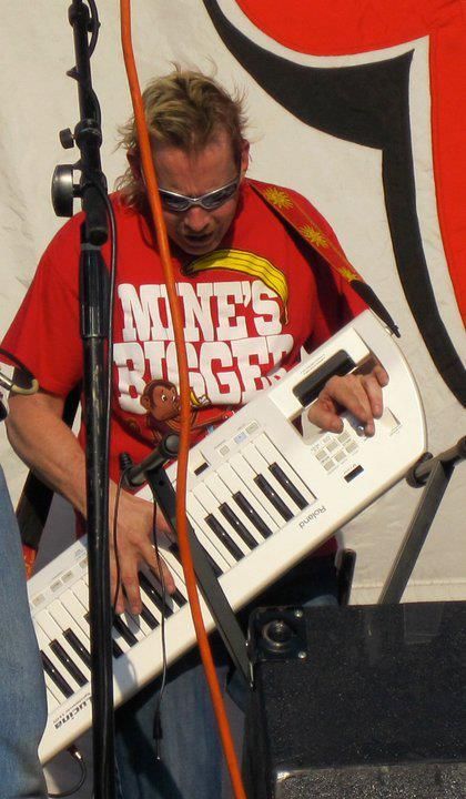 Man playing a white keytar on stage, wearing a red t-shirt that says 