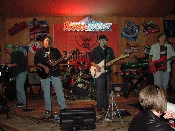 A band of five men performs on stage in a bar under the Coors Light sign. They are playing instruments with a mostly red and wooden background.