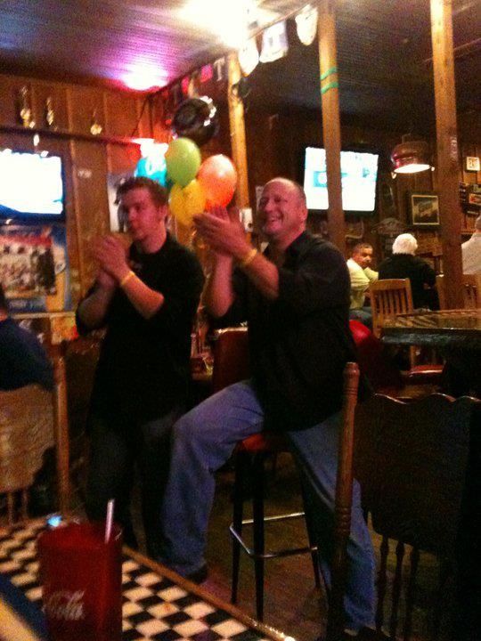 Two men clapping enthusiastically at a bar; one sits on a stool holding balloons, the other stands next to him.