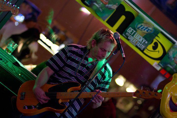 Man playing bass guitar in a bar. He's wearing a striped shirt, and there's a beer sign in the background.