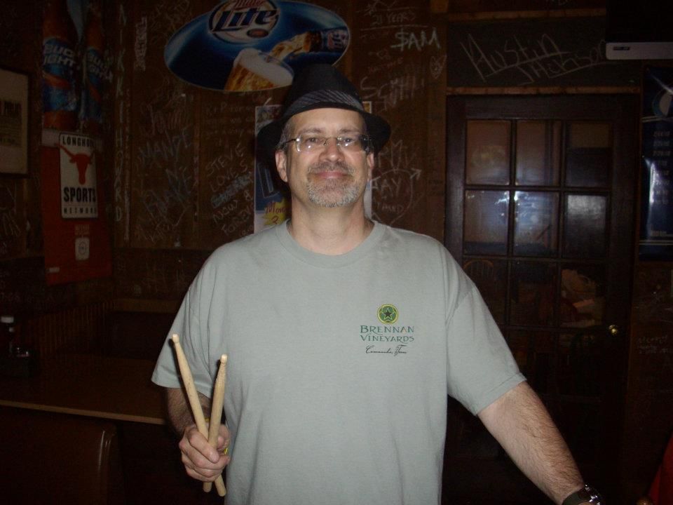 Man in hat and t-shirt holding drumsticks, standing in a bar. Brown walls and a Miller Lite sign in the background.