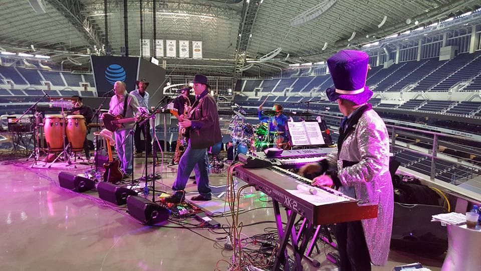 A band performs on a stage inside a large stadium. A keyboardist in a top hat and silver suit plays. Other musicians include a guitarist, drummer, and horn player.