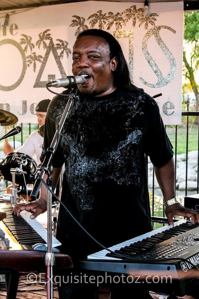 A black man with long hair sings into a microphone while playing a keyboard at an outdoor event, with a blurred background.