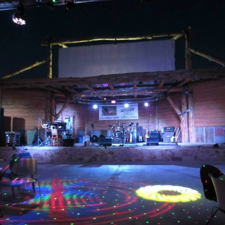 A band performing on a rustic stage with a screen, illuminated by stage lights and colorful floor projections.