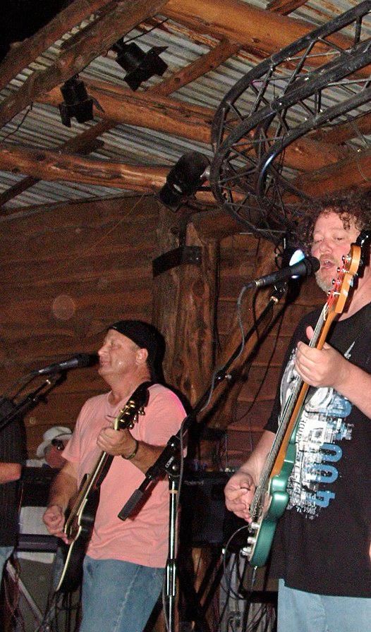 Band performing live on a wooden stage: two guitarists, a drummer partly visible, under stage lights.