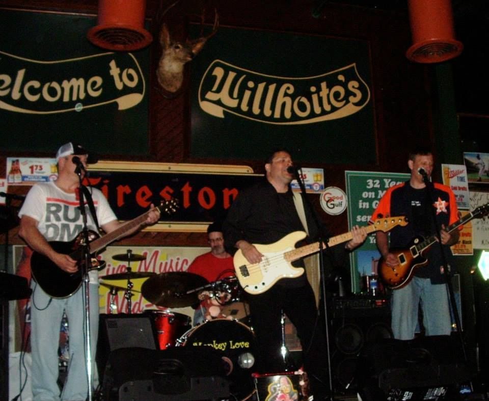 A band performs on stage at Willhoite's bar. Four men play guitars, bass, and drums under dim lighting.