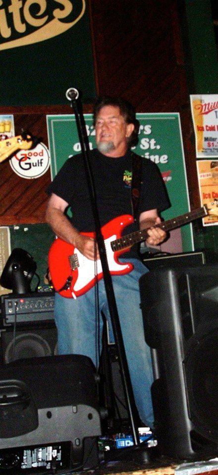 A man playing a red electric guitar on a stage. He's wearing a black shirt and jeans, with a microphone in front of him.