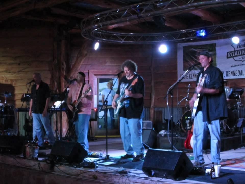 Band performing on stage: four men playing instruments, wooden interior, stage lights.