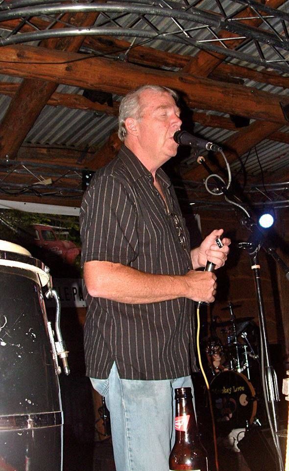 Man singing into a microphone on a stage, wearing a black striped shirt and jeans, with a drum set and other musical equipment visible in the background.