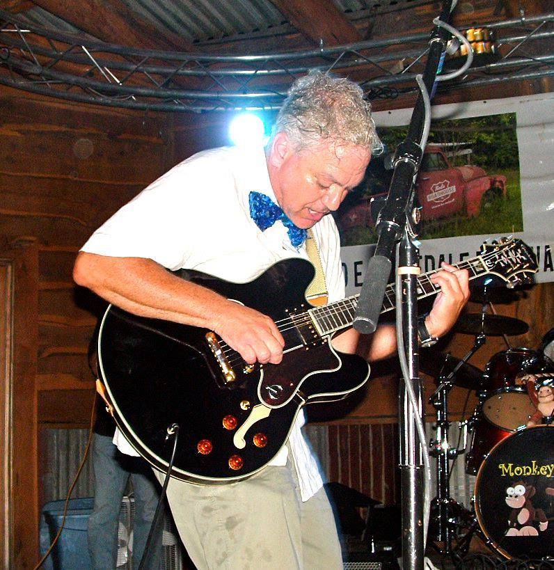 Man playing a black electric guitar on stage. He wears a white shirt, bow tie, and khaki pants. A drum set is in the background.