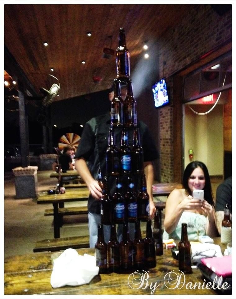 Man balancing a tower of beer bottles in front of a woman at an outdoor table. Dark setting, casual atmosphere.