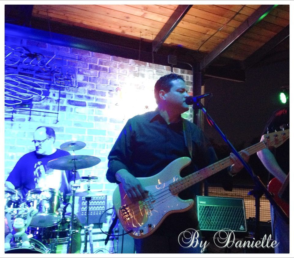 A band performing live on stage at a bar. A man plays a bass guitar and sings into a microphone, while a drummer plays in the background.