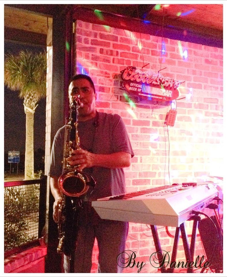 Man playing a saxophone and keyboard at a venue with red brick walls and colorful lighting.