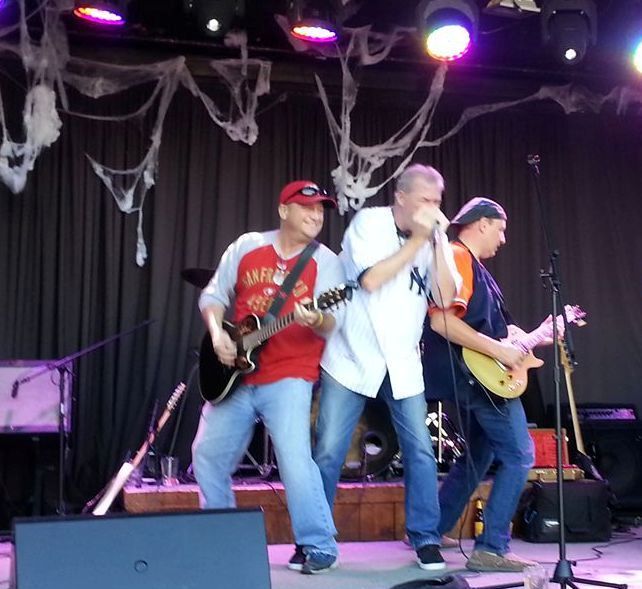 A band performs on stage: three men, one singing into a microphone, the others playing guitar. Stage is decorated with cobwebs, under colored stage lights.