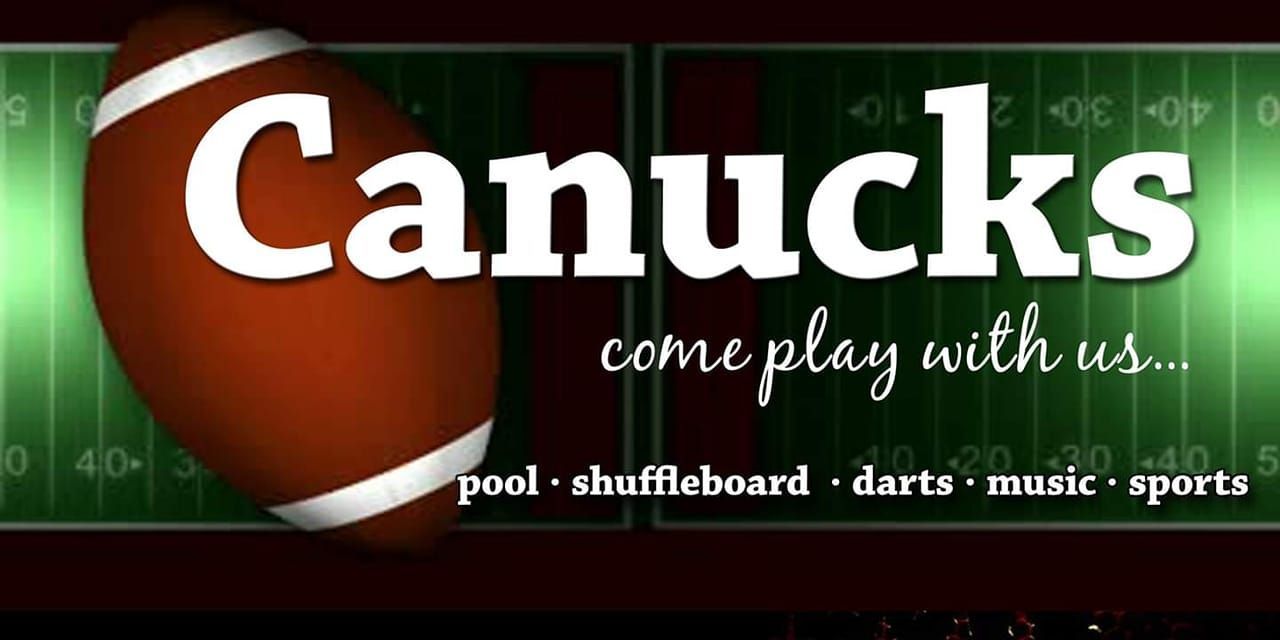 An outdoor restaurant patio at dusk. People sit at picnic tables under umbrellas strung with lights, near a large tree and a wooden fence.