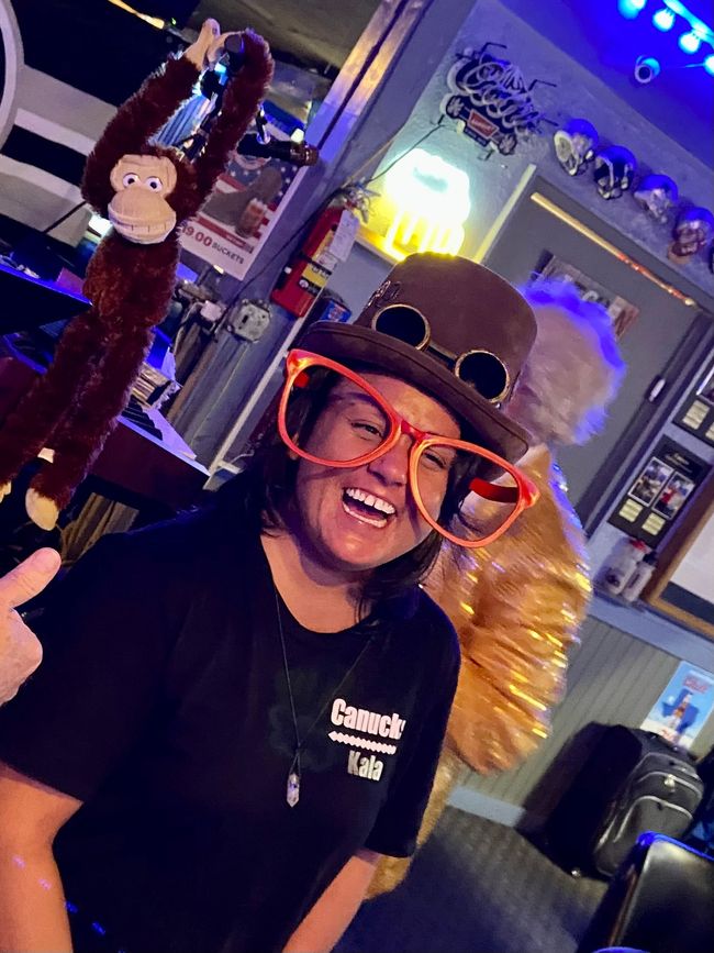 A woman wearing oversized orange glasses, a top hat, and goggles smiles widely. She is in a bar, a monkey decoration is beside her.