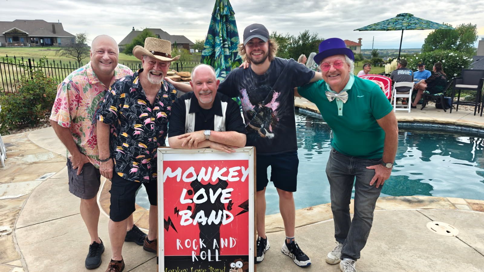 Monkey Love Band poses by pool. Five men: colorful shirts, holding a sign, smiles, sunglasses, background of houses.