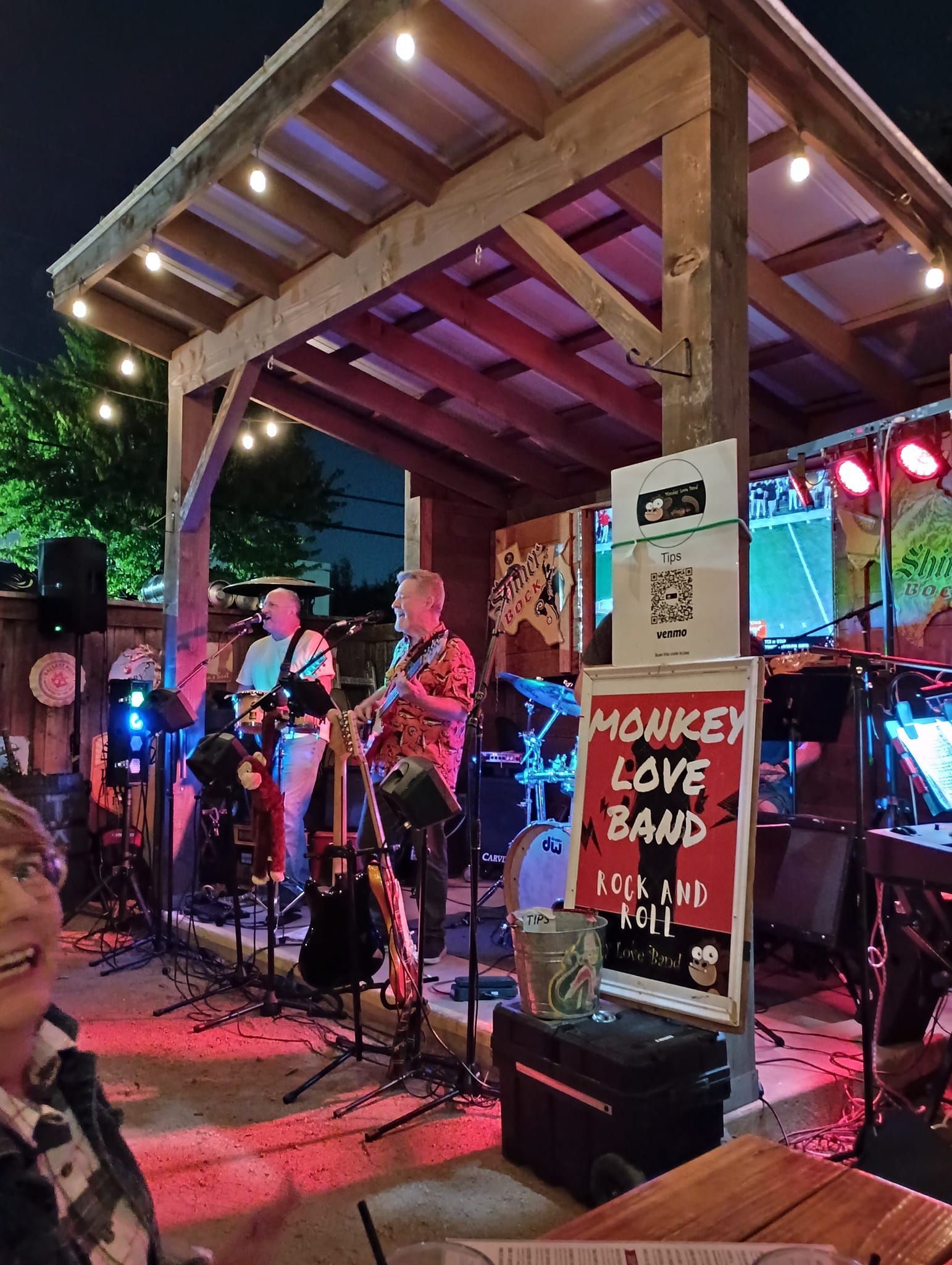 Band Monkey Love performs on a small outdoor stage with strings of lights. A woman in a plaid shirt is in the foreground.