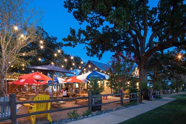 An outdoor restaurant patio at dusk. People sit at picnic tables under umbrellas strung with lights, near a large tree and a wooden fence.