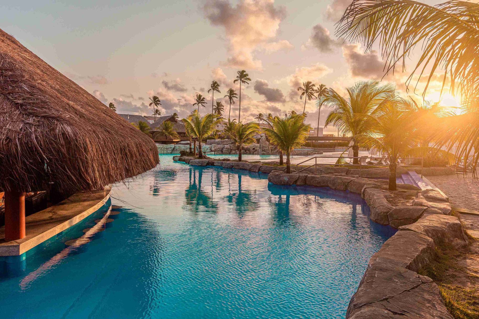 Vista do bar molhado sob quiosque de palha na piscina do resort Enotel Porto de Galinhas durante o pôr do sol, destacando a iluminação dourada e os coqueirais ao fundo