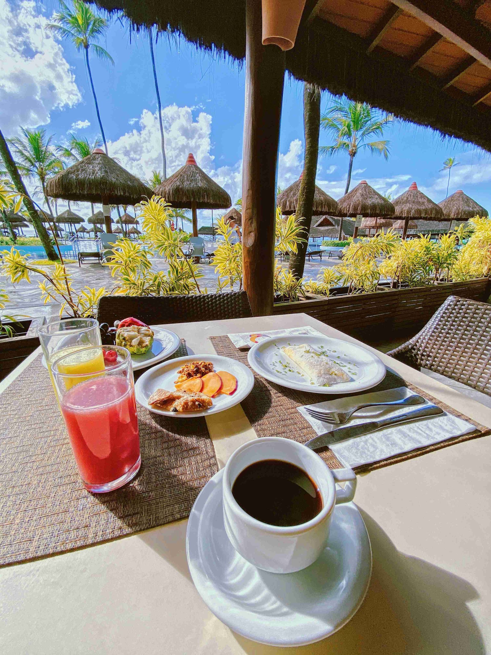 Mesa de café da manhã com café preto, tapioca, sucos naturais e frutas tropicais com vista para as piscinas