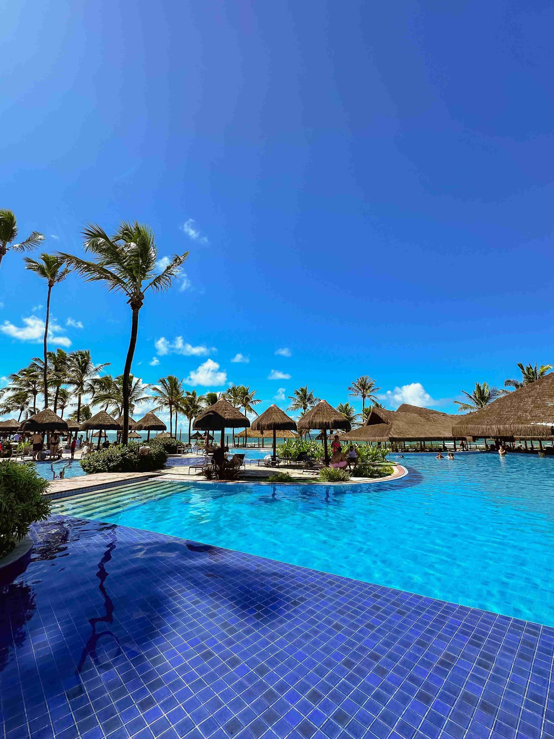 Piscina principal de azulejos azuis sob céu limpo, cercada por quiosques de palha e coqueiros no resort Enotel Porto de Galinhas